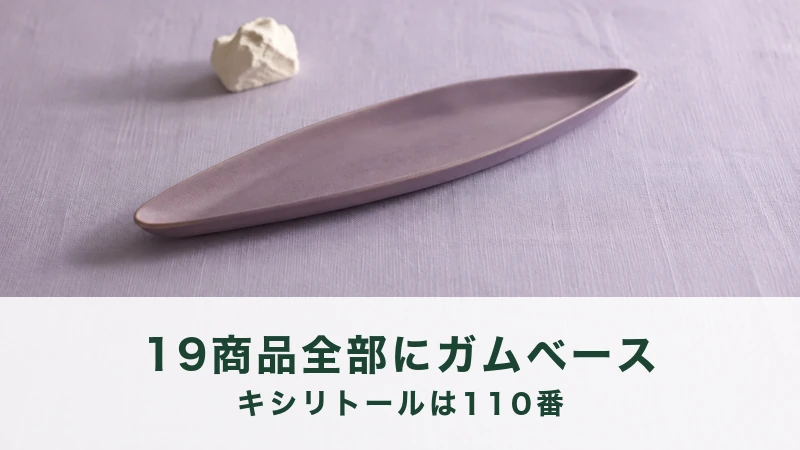 薄紫のリネンに藤色の細長い皿が斜めに置かれ、奥に生成りの石のかけらがぼける